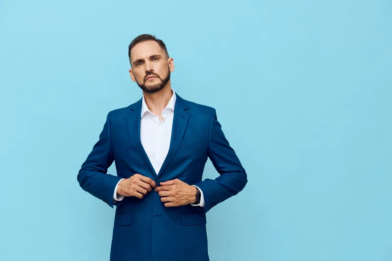 A professional man in a tailored blue suit stands against a solid blue background, projecting confidence, focus, and readiness for business matters.