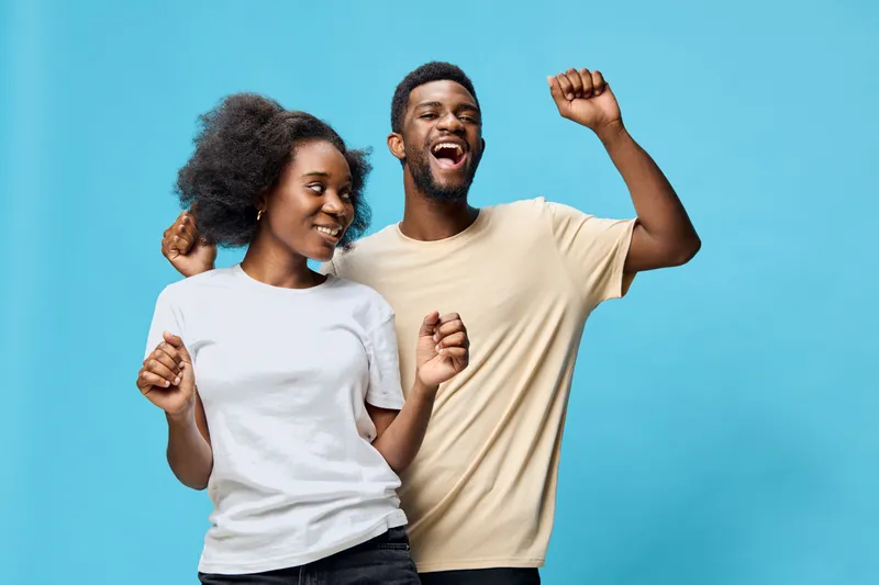 Happy young Black couple dancing joyfully on a vibrant blue background, wearing casual t shirts, expressing celebration and excitement together