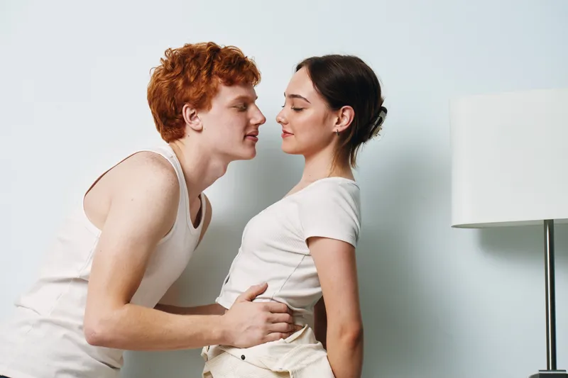 Young couple standing close indoors, man leaning in to kiss woman, casual clothes and relaxed mood, modern room with lamp and neutral wall background, intimacy and affection.