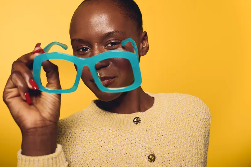 Close-up of confident African woman with dark skin holding large blue glasses in front of her face against yellow background. Concept of fashion, style, and beauty portrait.