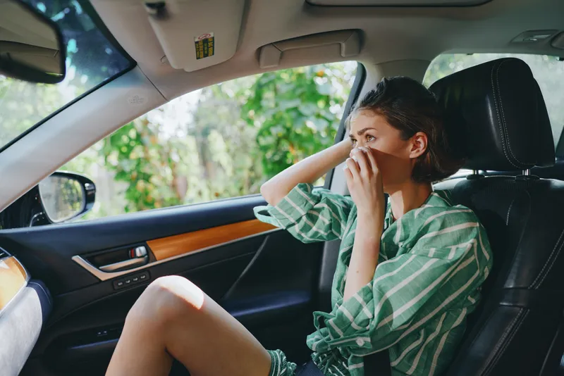 A woman sits in the driver's seat of a car, relaxed and thoughtful, wearing a green striped shirt, looking ahead.