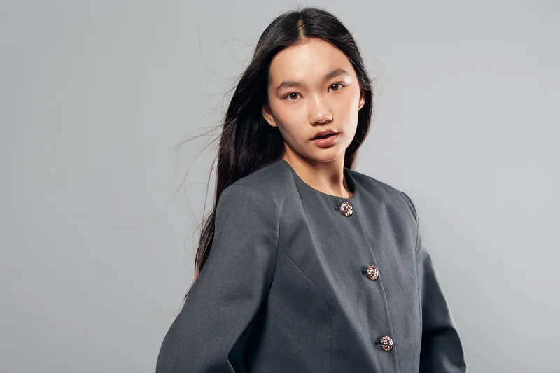 Young woman with long black hair wearing gray buttoned jacket posing against light gray background for fashion portrait and style concept with natural expression and modern clothing.