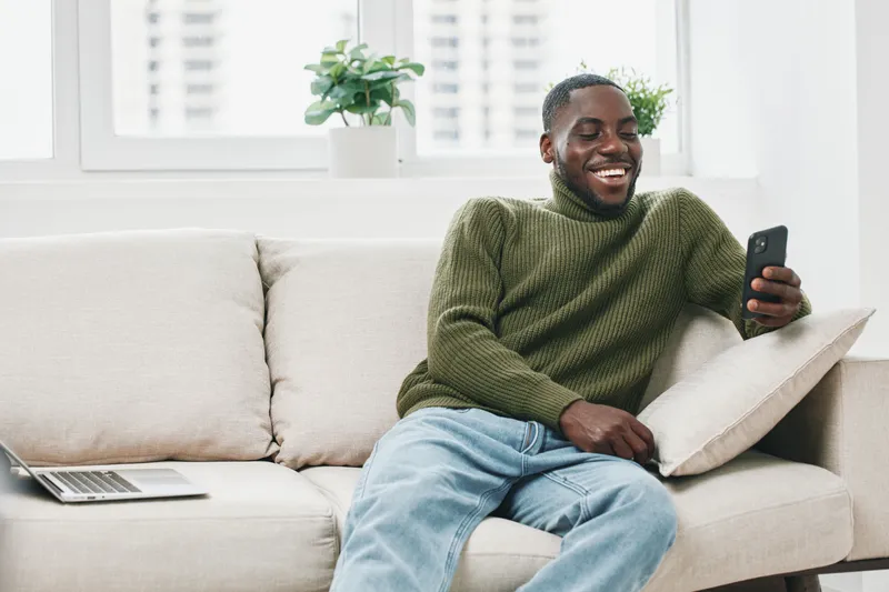 Smiling young Black man relaxing on a sofa, using a smartphone in a cozy, modern living room, conveying joy and comfort