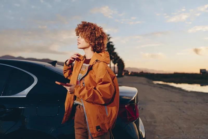 Woman lifestyle by car on road at sunset, curly hair, orange jacket, holding a snack near the vehicle, relaxed travel mood, warm sky and distant hills, outdoors with soft smile and freedom