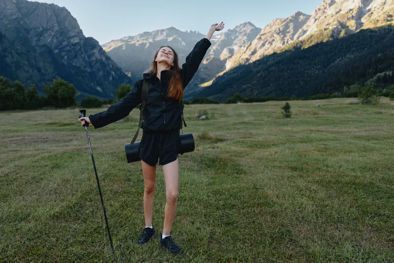 A joyful outdoor explorer stands in a grassy field with mountains in the background, arms raised and head tilted back, capturing a sense of freedom and adventure.