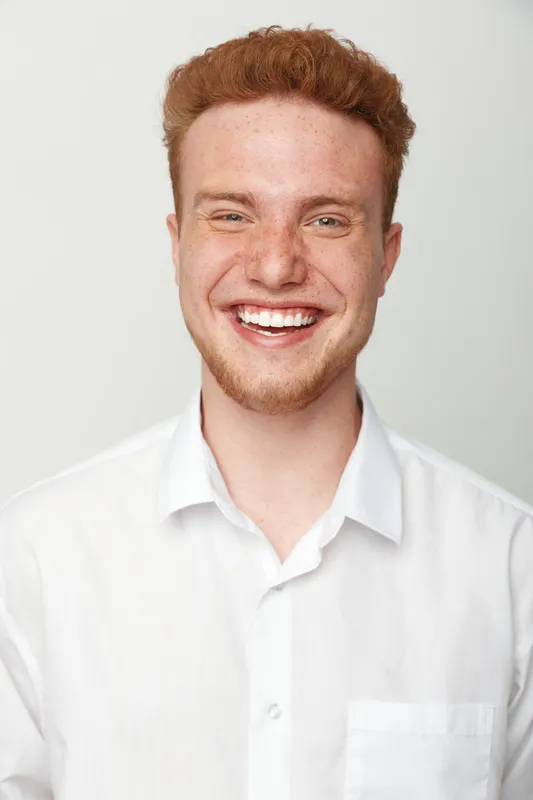 Smiling young man with red hair and freckles wears white shirt, looking at camera on light background. Concept of happiness, youth, and natural beauty in portrait photography.