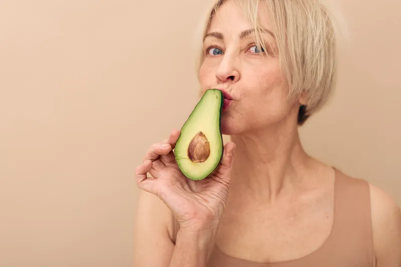 Mature woman with short blonde hair holds and kisses fresh avocado half. Neutral background with natural light emphasizes healthy lifestyle and organic food concept.