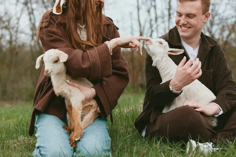 A couple with goats and kids share a tender outdoors moment in a grassy field, smiling and interacting with the animals, capturing a warm lifestyle scene of countryside companionship and farm life