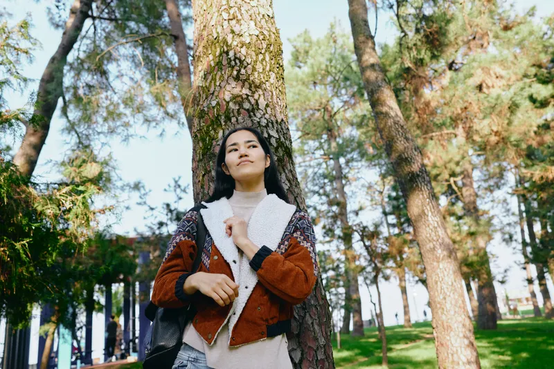 Forest setting with a stylish woman wearing a warm coat, standing among tall trees as sunlight filters through, capturing nature, fashion and a calm outdoor moment