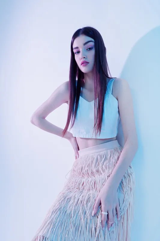 Young woman posing in a white crop top and textured skirt. Her long hair is straight, and she looks confidently at the camera. Studio shot with soft blue lighting. Concept of woman beauty fashion.