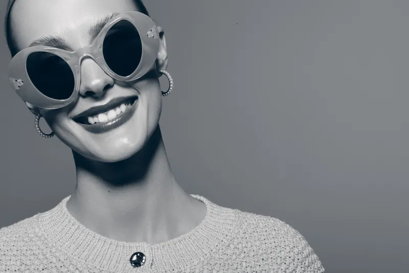 Smiling young white woman with pale skin wearing large round sunglasses and earrings looks happy in studio with neutral background and knitted sweater.