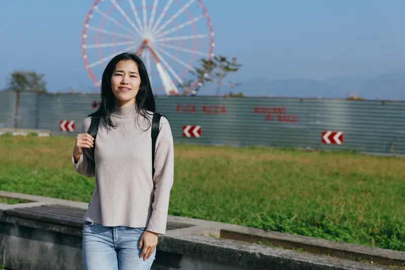Traveler woman stands beside a railing in a sunny outdoor setting with a distant ferris wheel, backpack slung, wearing a light sweater and jeans, capturing a casual travel mood in a park like scene
