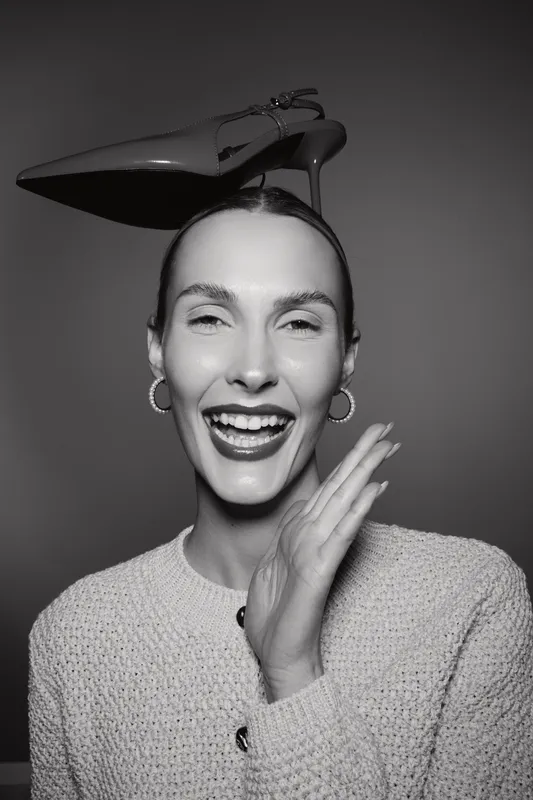 Smiling Caucasian woman with light skin wearing hoop earrings and a knitted sweater balances a high heel shoe on her head. Portrait in black and white with joyful expression.