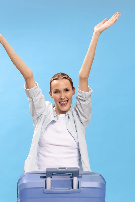 Happy woman with raised arms stands behind suitcase on blue background. Concept of travel, vacation, tourism, and joy. Casual clothing and smiling face express excitement and freedom.