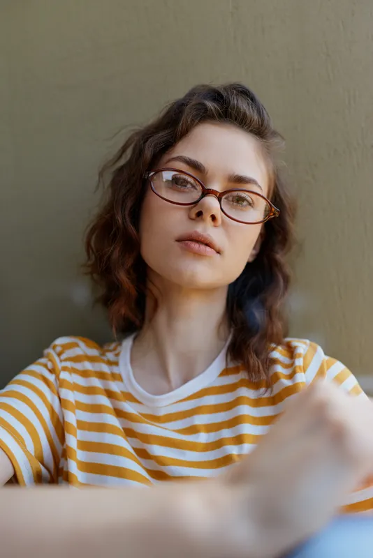 A young woman with wavy brunette hair and glasses sits against a neutral wall in a striped yellow and white shirt, looking at the camera with a calm, thoughtful expression.