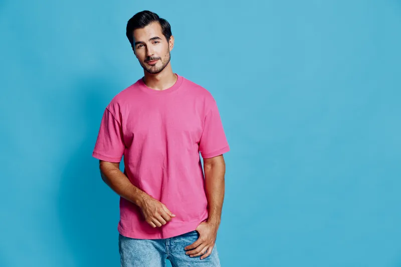 Smiling young man in a pink t-shirt poses confidently against a solid blue background, showcasing a casual and relaxed vibe. People lifestyle concept.