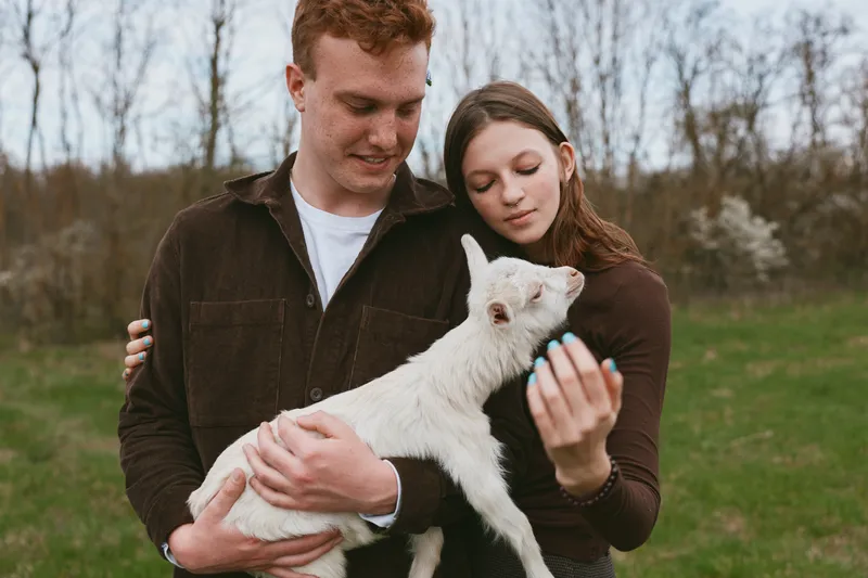 Couple outdoors with a calm goat kid, holding the animal and sharing affection in a sunny field setting, natural portrait of rural life and gentle bonding