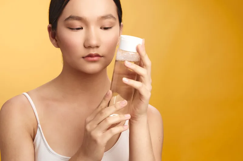 Asian woman in a bright studio examines skincare, holding a cosmetic jar calmly. She seems focused and at ease, conveying a quiet moment of self care and confidence.
