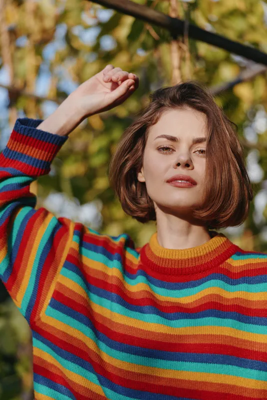 Woman in her late twenties with bob haircut wearing a rainbow sweater, outdoor portrait raising her arm and smiling confidently among autumn trees in warm natural light.
