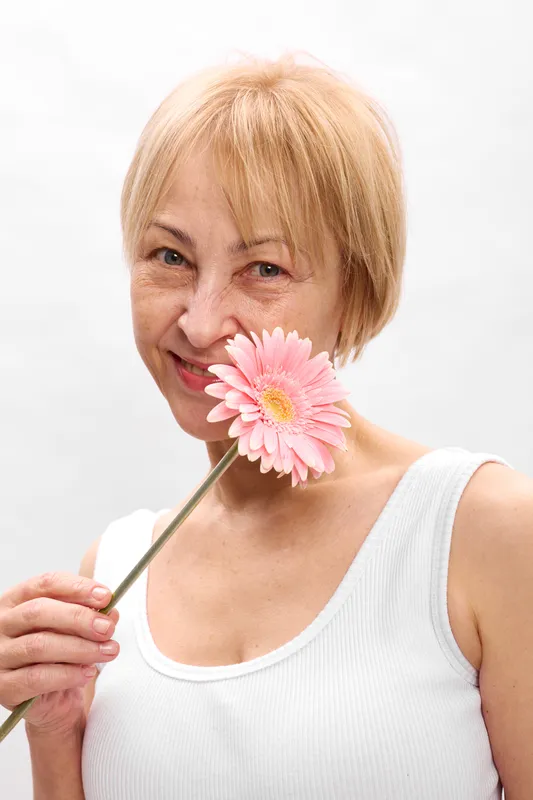 Smiling woman, flower near lips, portrait closeup in studio, blonde short hair, pink blossom, soft light on white background, relaxed gaze, peaceful spring mood