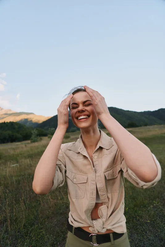 Joyful woman outdoors, laughing with hands in her hair, casual beige shirt, sunlit moment, natural colors, lush greenery background, carefree lifestyle in open meadow