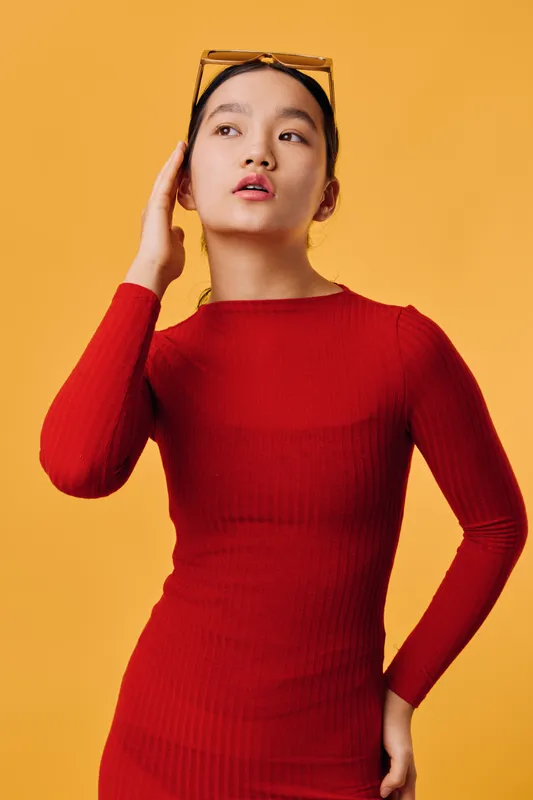 Young woman posing in red long sleeve dress against yellow background. Fashion model with sunglasses on head looking aside, confident and stylish in studio portrait.