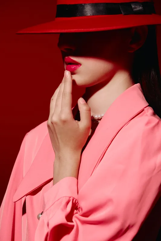 Fashion model in neon pink jacket and red hat poses with finger near bright red lips in high-contrast studio lighting. Concept of beauty and creative fashion style.
