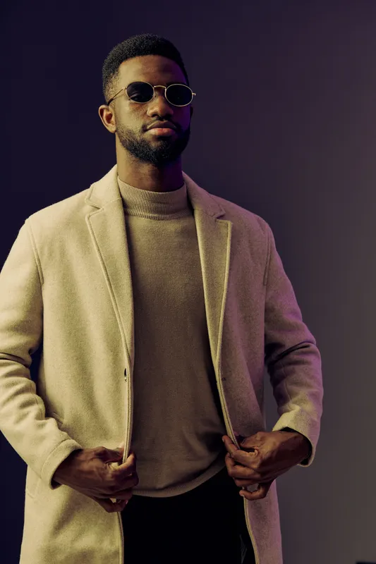 Stylish young man in a beige coat and sunglasses, exuding confidence against a modern purple backdrop, perfect for fashion related themes, urban lifestyle, and seasonal trends