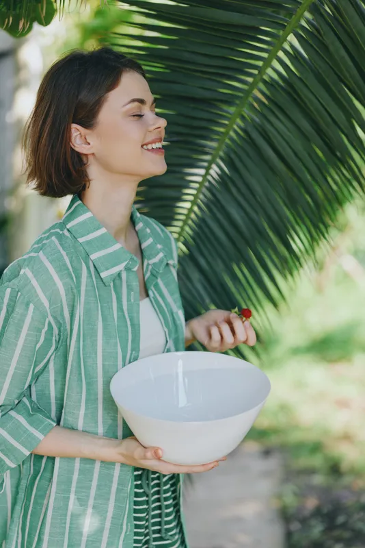 A cheerful woman outdoors holds a white bowl, smiling warmly under dappled sunlight and lush palm leaves.