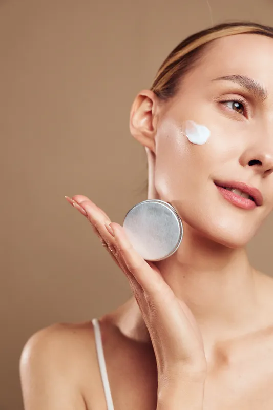 A young woman applies white facial cream on her cheek with a silver container in hand. The neutral background highlights skincare, beauty, and daily self-care routine concepts.