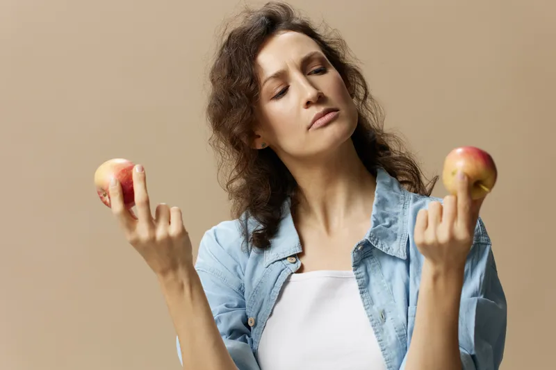 Задумчивая, сомневающаяся, кудрявая, красивая женщина в джинсах и повседневной рубашке смотрит на яблоко, позирует изолированно на бежевом пастельном фоне