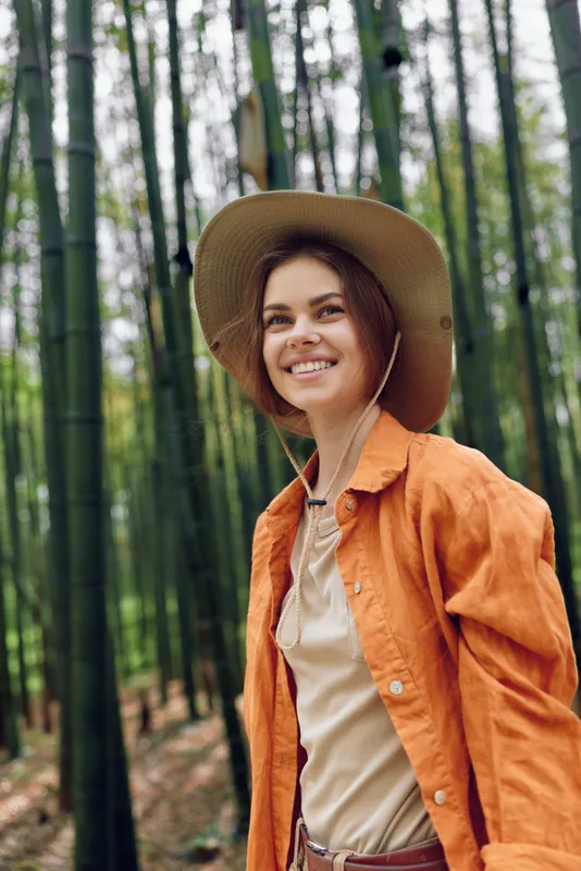 Woman smile in bamboo forest wearing wide hat and orange jacket, portrait in nature outdoors. Young traveler enjoying green grove, happy expression and casual autumn outfit