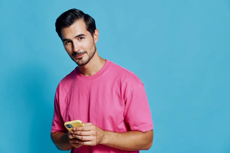 Stylish man smiling while holding a yellow mobile phone in his hand against a bright blue background. Casual wear and modern vibe emphasize his youthful energy.