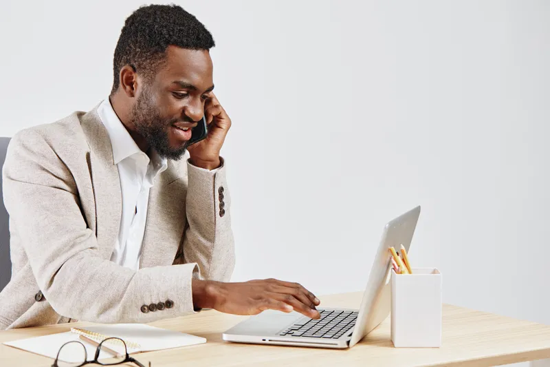 Confident young African American man smiling while talking on phone and working on laptop in bright office setting. Casual business style, computer, glasses, modern workspace, technology, communicatio