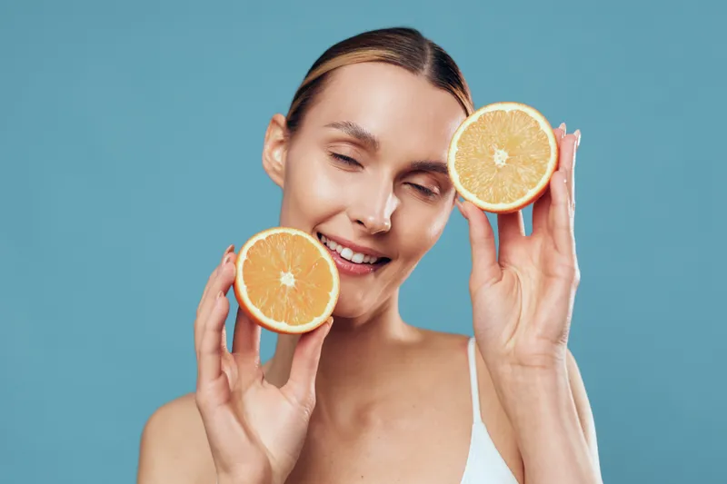 Smiling woman holding two halves of a fresh orange near her face on blue background. Concept of healthy lifestyle, vitamin C, and natural beauty. Soft lighting highlights fresh fruit and skin.