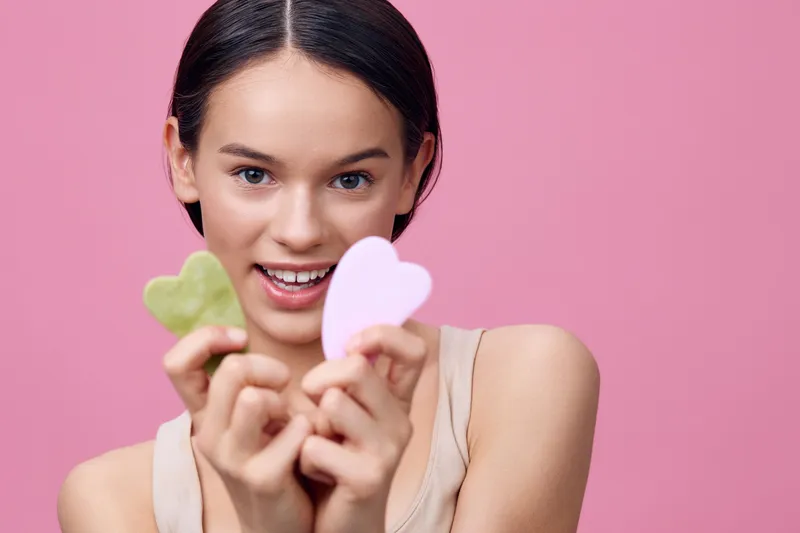 Young woman holding heart shaped beauty tools, showcasing skincare and self care solutions in a vibrant pink background, promoting healthy beauty practices