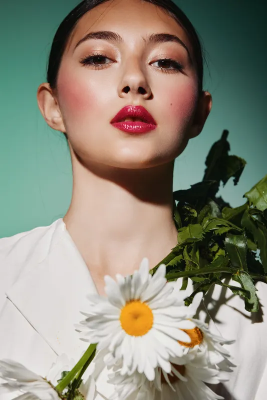 Close-up portrait of a fashionable young woman with shining wet-look lipstick and floral greens against a soft green background. Beauty creative look highlighting makeup and natural elements.