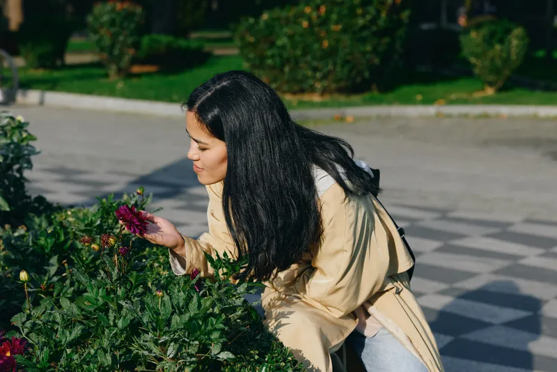 A woman bends toward flowering bushes outdoors, smelling blossoms in a garden setting, capturing a serene moment of nature, fragrance, and gentle outdoor activity.