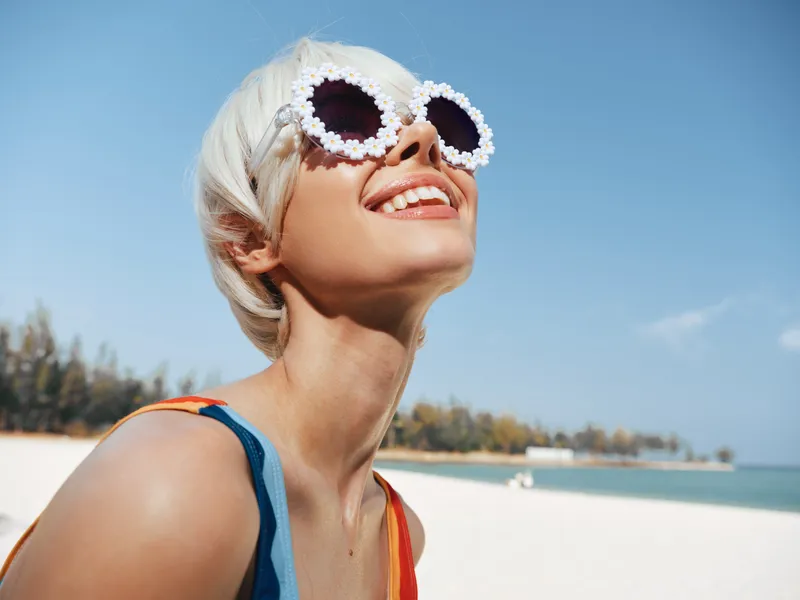Joyful portrait of a smiling woman with short white hair wearing flower-framed sunglasses, basking in bright sun on a sandy beach under a clear blue sky.