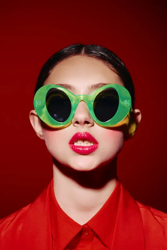Young woman wearing large green sunglasses with dark lenses poses against a bold red background. She wears a bright red blouse and glossy red lipstick. Beauty and fashion concept.