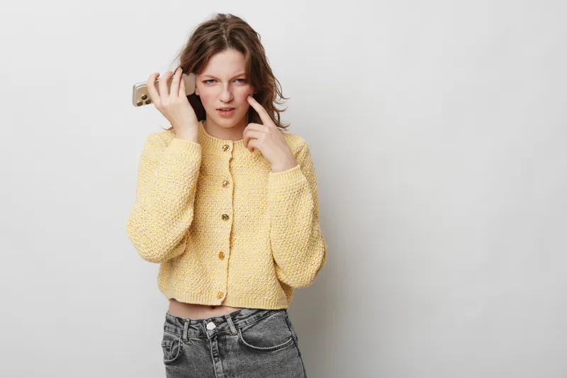 Confused young woman in yellow knitted sweater holds smartphone to ear and points to face, expressing doubt or misunderstanding. Isolated on white background with casual outfit and natural light.