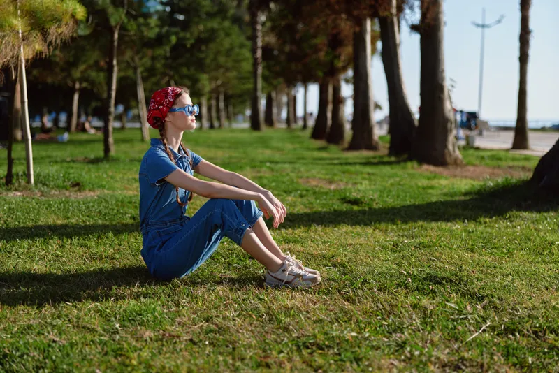 Lifestyle woman in modern boho-western denim street style sitting on grass, wearing sunglasses and floral headband, captured in warm film color tones outdoors.