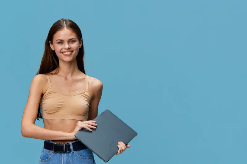 smiling young woman holding laptop, casual outfit, blue background, happy expression, technology, lifestyle concept