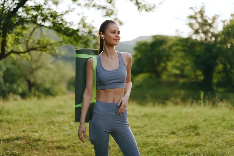 Young athletic woman in workout clothes with yoga mat, smiling in a green outdoor setting, promoting fitness and healthy lifestyle