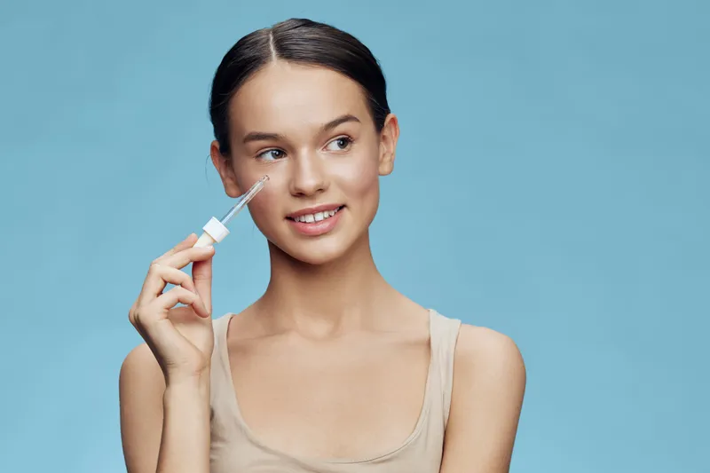 Young woman applying serum for skincare with a bright blue background, showcasing a healthy complexion and a joyful expression, representing beauty and self care