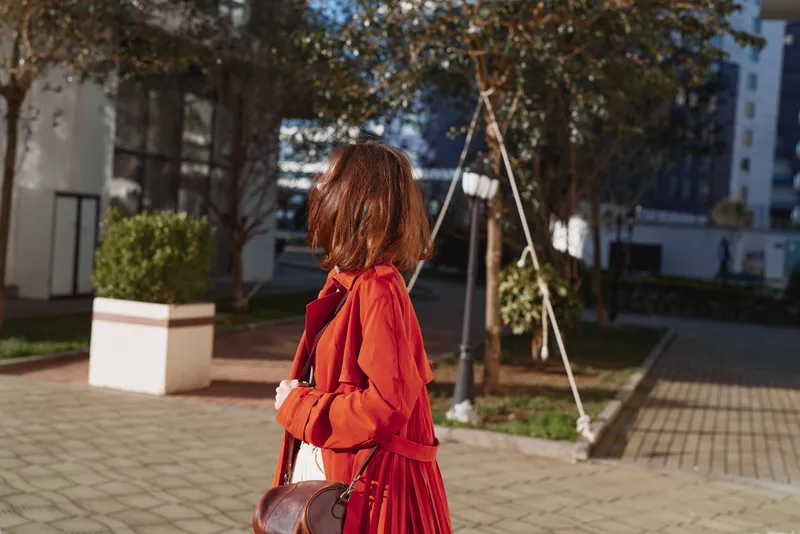 An adult woman in a red dress walks along a sunlit urban street, carrying a brown crossbody bag, captured in candid fashion for a lifestyle moment.