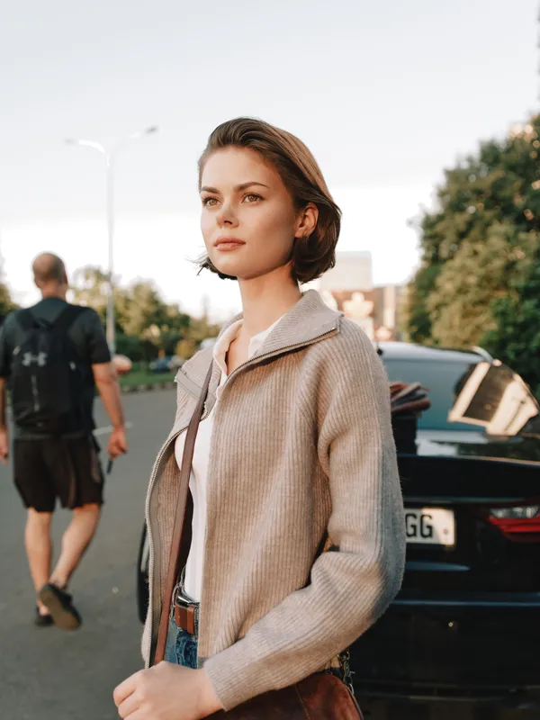 A poised young woman stands outdoors near a parked car, looking into the distance with calm confidence. The scene conveys urban style and independence, capturing a candid street portrait with