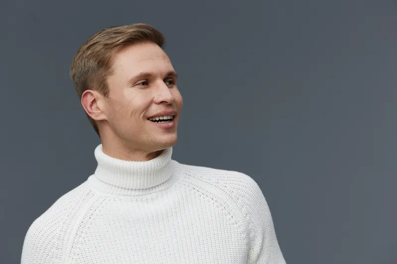 Smiling man wearing white turtleneck sweater looking to the side on plain gray background studio portrait. People lifestyle concept, positive expression, casual style, warm clothing, relaxed mood, hor
