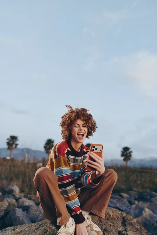 Smiling woman with curly hair holding a smartphone for a selfie outdoors on rocky terrain. Lifestyle moment in nature, mountains and blue sky in background, travel mood, casual clothing.