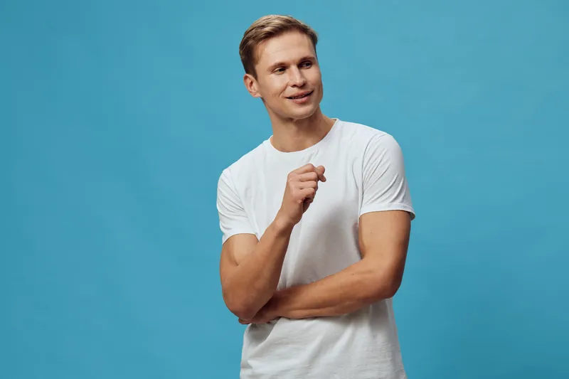 Smiling young man with light brown hair wearing casual white t-shirt, posing confidently with crossed arms on a plain blue background. Studio portrait of a cheerful male model, minimalistic style, lif